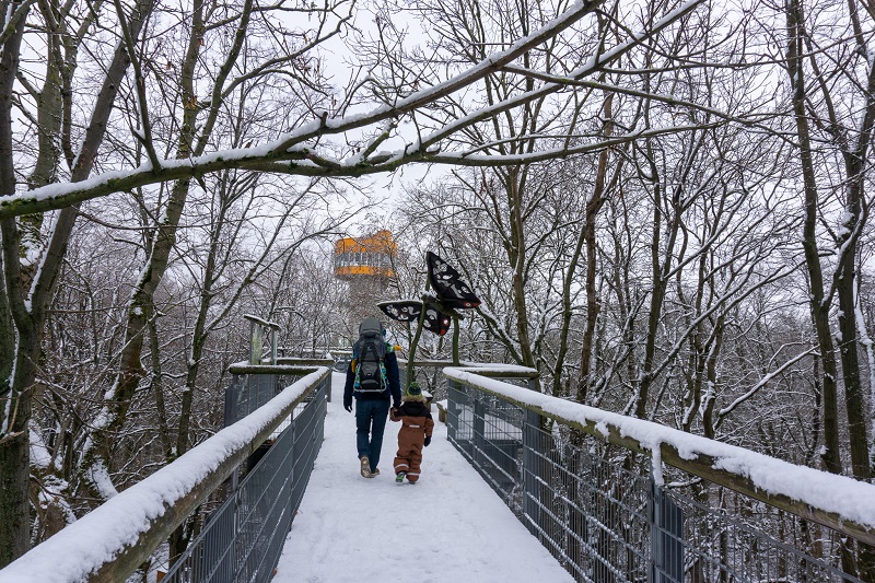 Ein Vater läuft mit seinem Kind an der Hand über den verschneiten Baumkronenpfad im Nationalpark Hainich. Vor Ihnen ist ein Holz-Schmetterling und der orangefarbene Aussichtsturm zu sehen.
