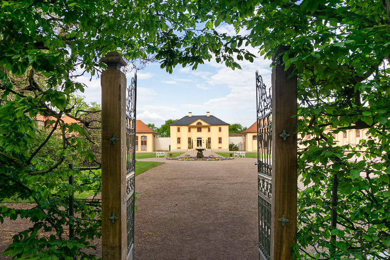 Blick durch eine Hecke mit geöffnetem eisernen Tor auf das Hauptgebäude der Orangerie des Schlosspark Belvedere.