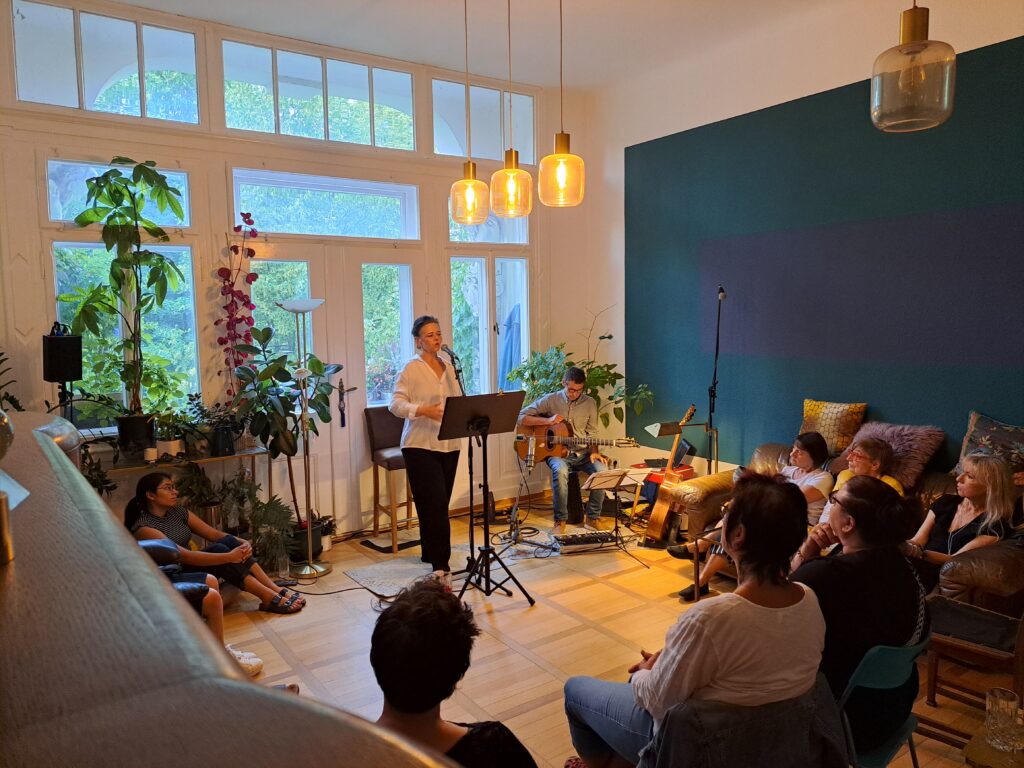 Sandra Peschke steht hinter ihrem Mikrofon im Wohnzimmer und singt. Neben ihr spiel Marco Böttger die Gitarre.