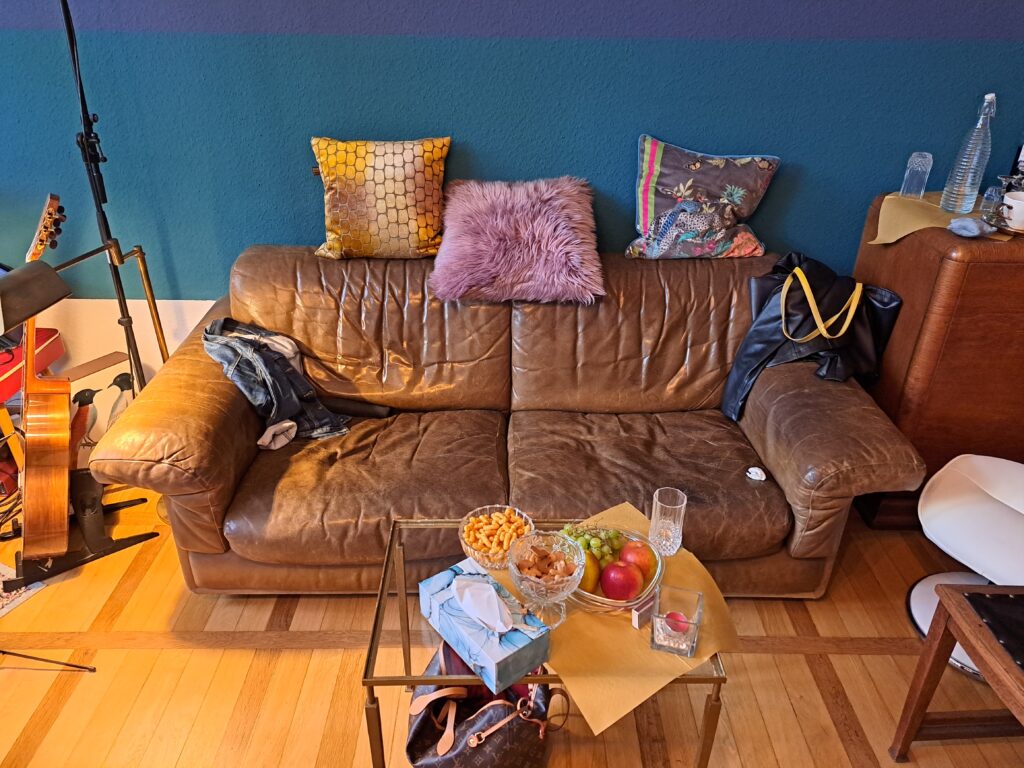 Eine braune Ledercouch, geschmückt mit bunten Kissen, steht vor der blau-grünen Wand im BlauGrünen Salon.